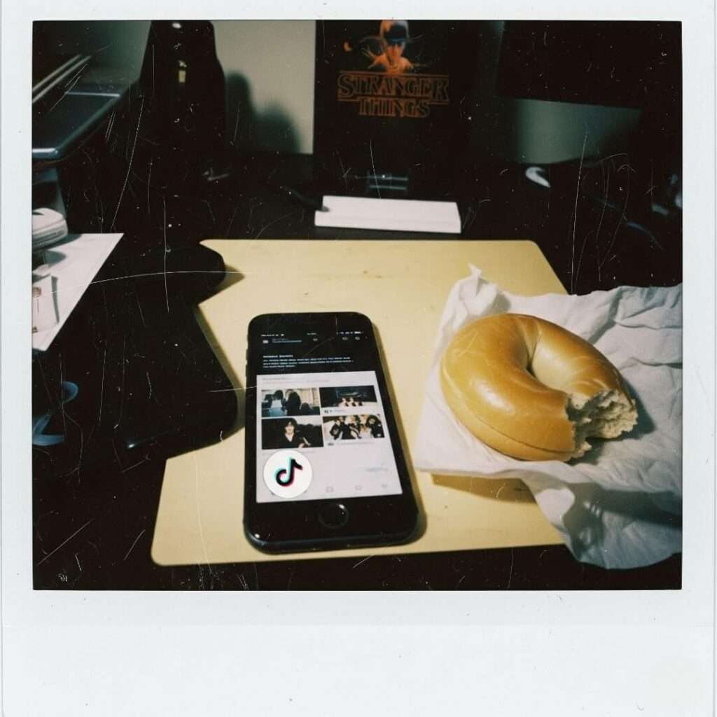 A vintage Polaroid photo showing a messy desk with a smartphone open to TikTok, a half-eaten bagel, and a "Stranger Things" poster in the background. A musical note emoji sticker is on the phone's screen.